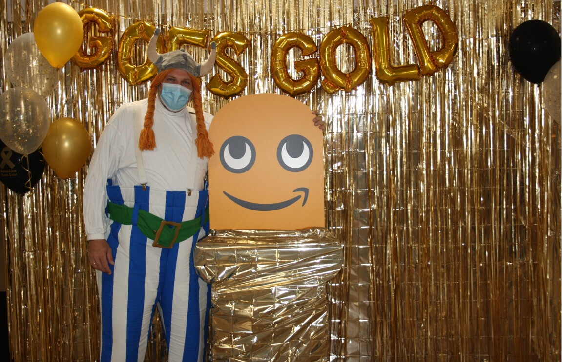 Ein verkleideter Mitarbeiter steht vor einer "Amazon Goes Gold"-Wand.