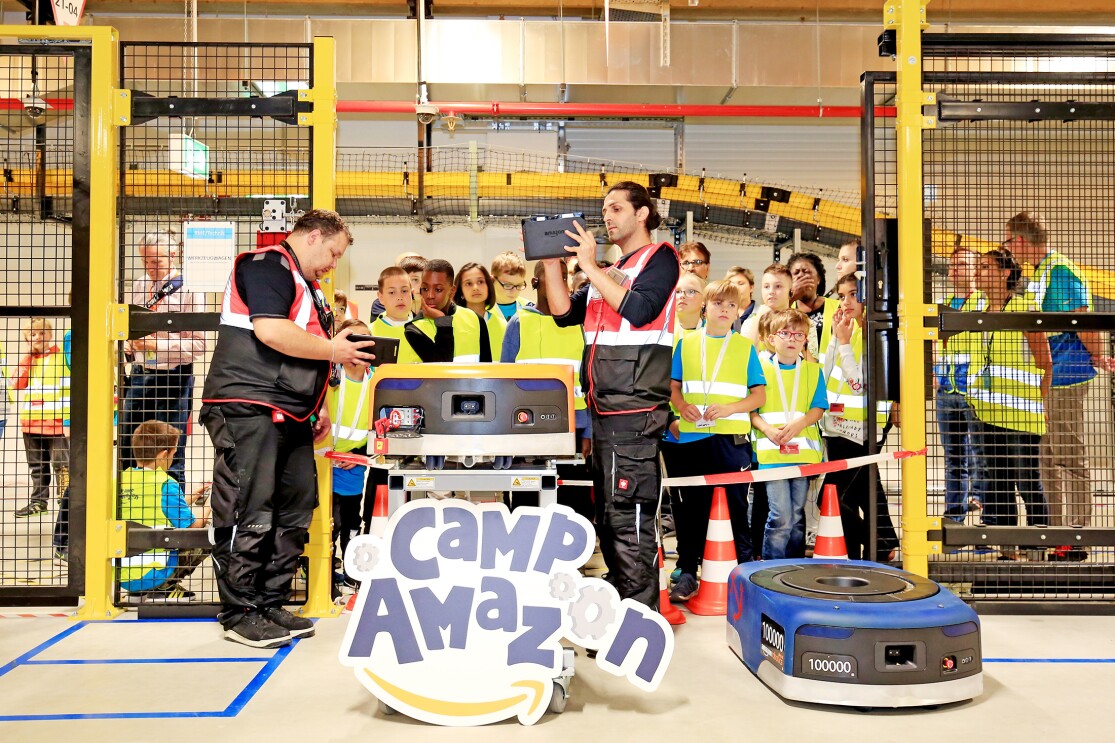 Eine Gruppe von Kindern hat sich in einer Logistikhalle rund um einen Transportroboter versammelt, der auf einem Tisch steht. Zwei Amazon Mitarbeiter erklären ihnen die Funktionsweise.