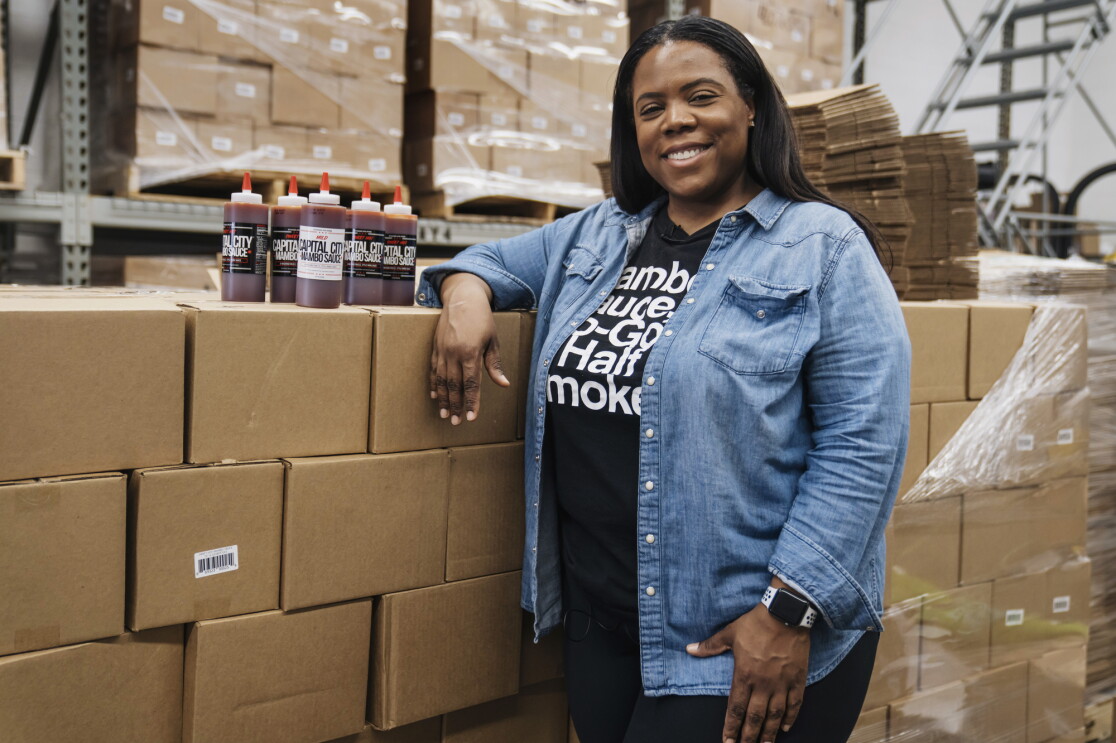 Arsha Jones smiles as she stands in front of stacks of boxes with a handful of Capital City Mambo Sauces next to her on top of the boxes.