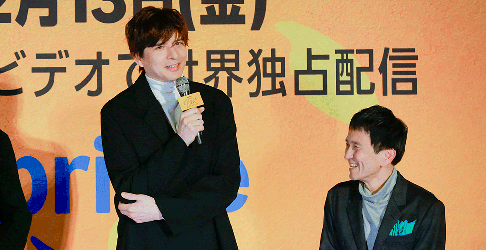 オレンジ色の壁の前、オレンジのステージの上で並ぶ2人の人物。左側の背が高い男性がマイクを持って話している。