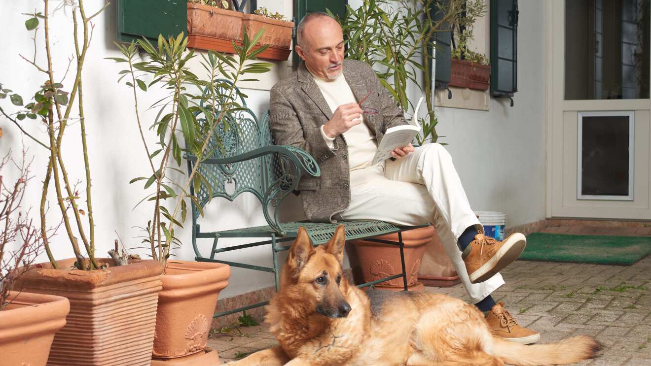 Lo scrittore Federico Maria Rivalta legge un libro sul portico di casa con un pastore tedesco ai suoi piedi.