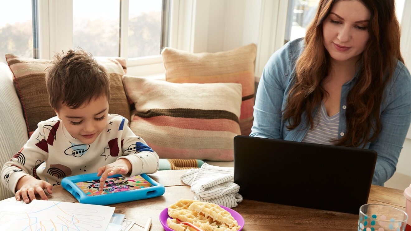 A mother sits alongisde her son at the breakfast table. She works on her laptop while her son plays on his Kindle.