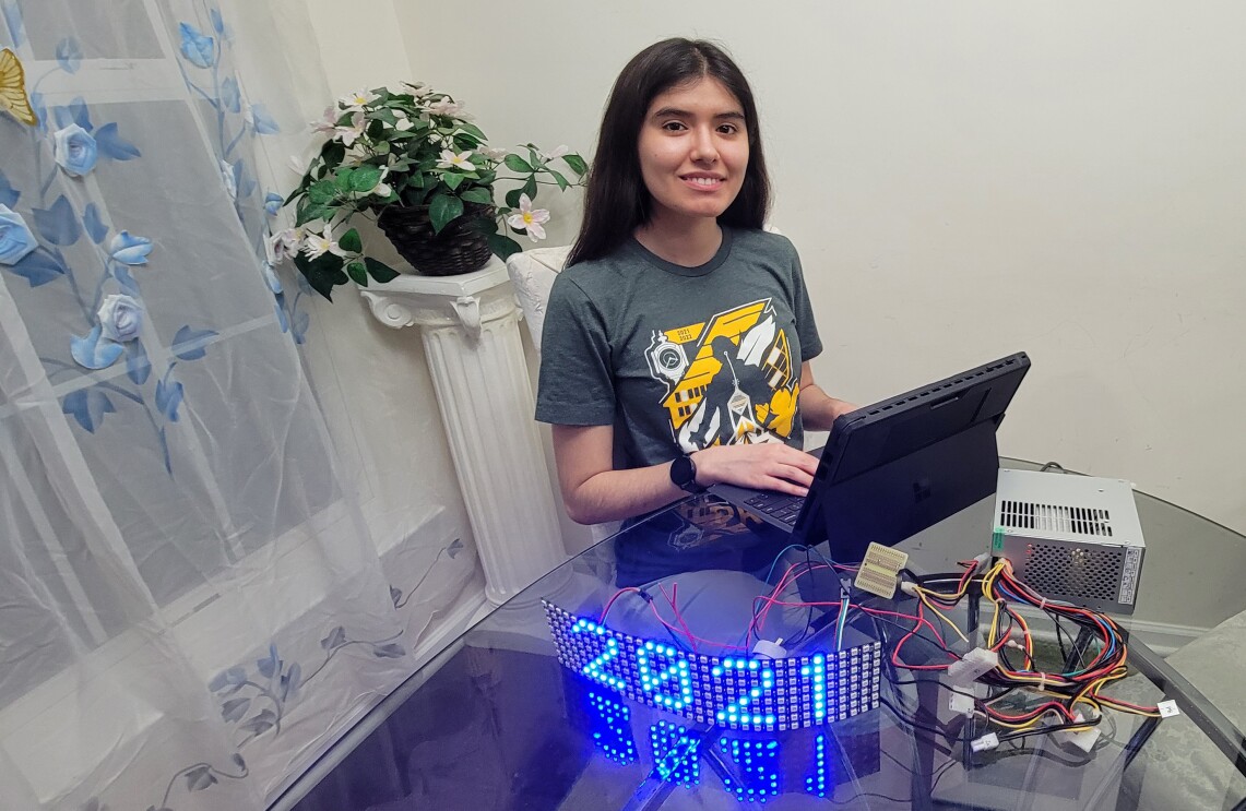 An image of a young woman sitting at a dining room table smiling for a photo. In front of her is a laptop hooked up to wires and an illuminated sign that says "2021."