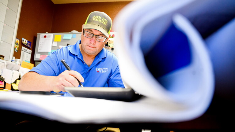 A photo of the owner of a Fence company working at a desk, holding a pen. There are a scroll of drawings in front of him.