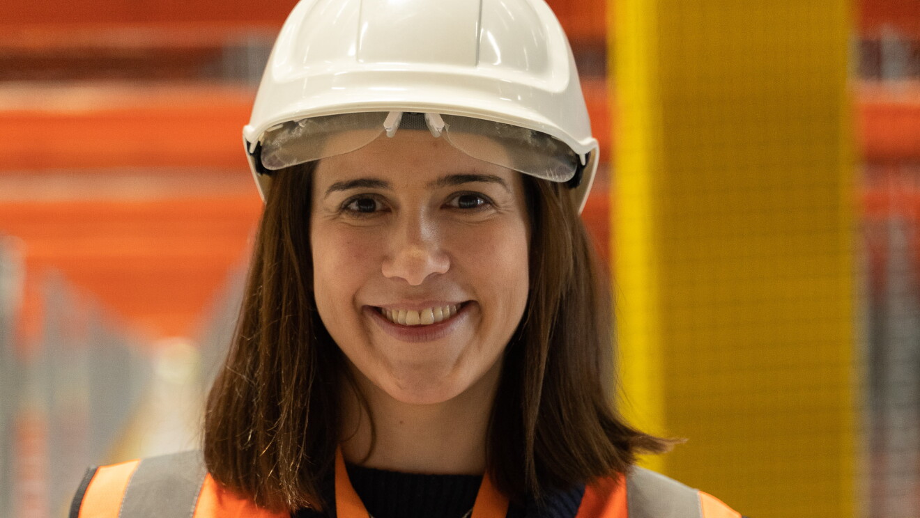 Isabel Bermúdez, Responsable Senior de Proyectos de Ingeniería para Amazon España, mirando a cámara y sonriendo. Lleva casco blanco, chaleco naranja. Tiene el pelo por el hombro. El fondo está difuminado.