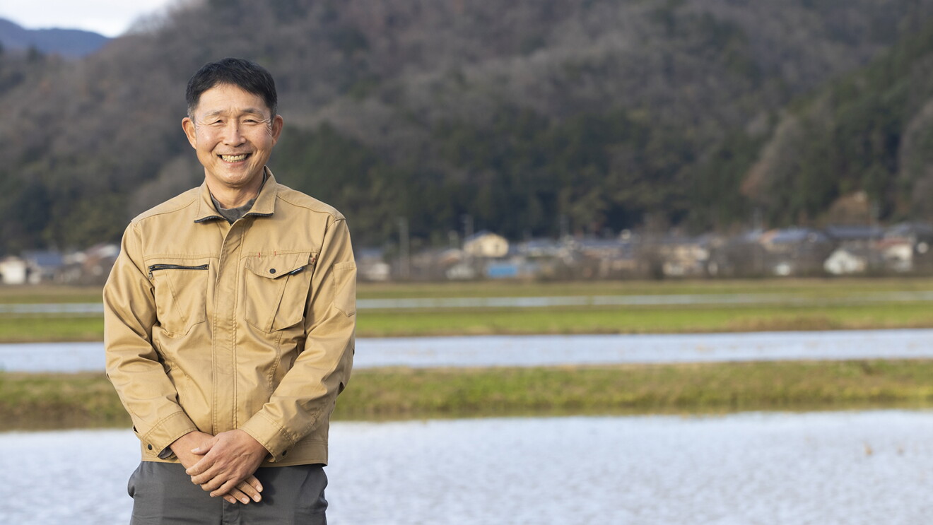 美しい里山と水田を背に立つやわらかな表情でほほ笑む男性