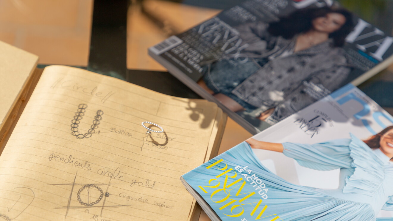 SOnia Llamas de Iyé Biyé está preparando una nueva colección. En la foto hay dos revistas de moda y una libreta con sus diseños dibujados. Al lado de uno de sus diseños, la joyas hecha realidad. 