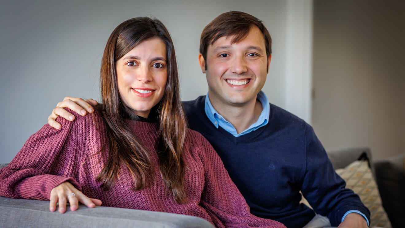 Miriam y Alberto sentados en el sofá de su casa. El sofá es de color gris, la pared también y encima del sofá un cojín estampados con colores blancos, grises y amarillos. Los dos están mirando a cámara. Ella va con un jersey de punto morado, el pelo largo castaño hasta el pecho y tiene su mano izquierda apoyada en la pierna de Alberto. La otra mano la está apoyando en el apoyabrazos del sofá. Él la agarra por el hombro con la mano derecha. Lleva un pantalón de color beige, un jersey azul oscuro y una camisa azul clarito. 
