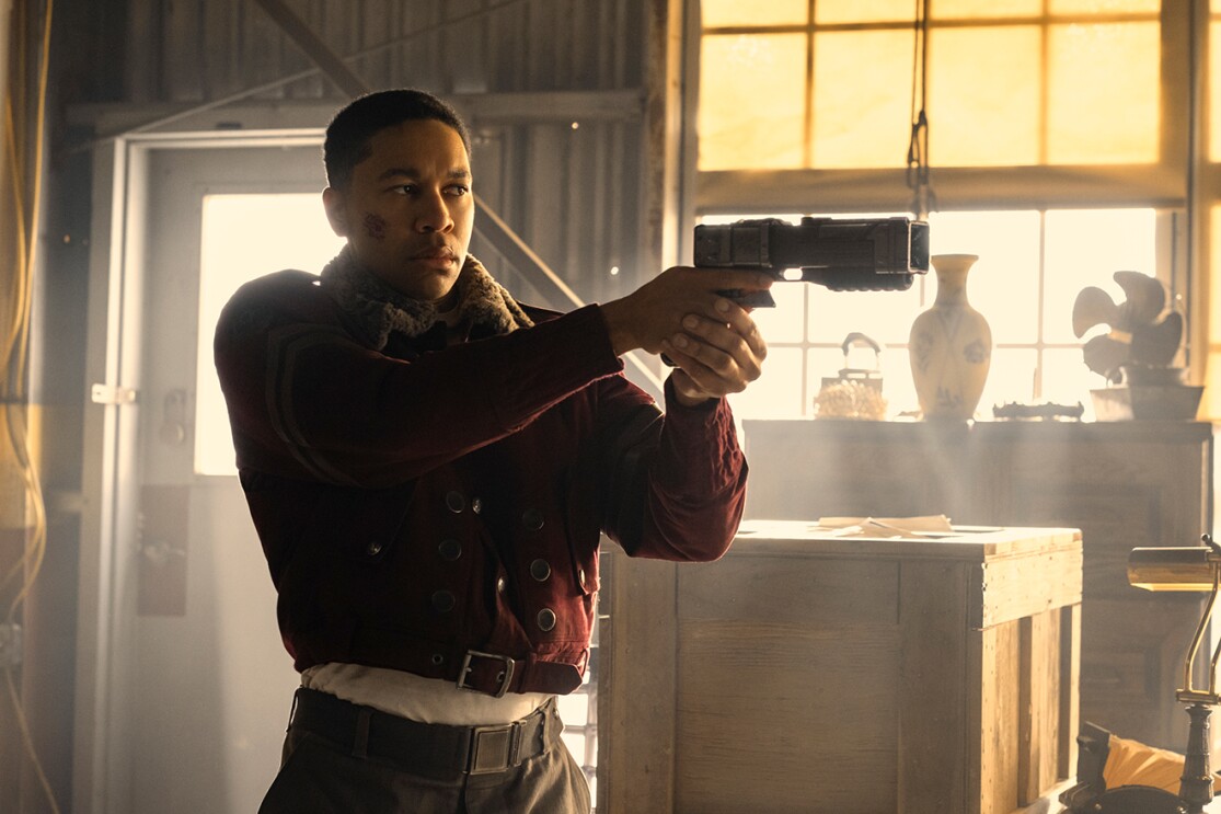 窓から黄色い光が差すほこりっぽい室内に立つ、赤いジャケットの男性。未来的な銃を両手で構え右側に向けている。窓の前に壺や古い扇風機などが並ぶ。男性の背後に木製クレート。