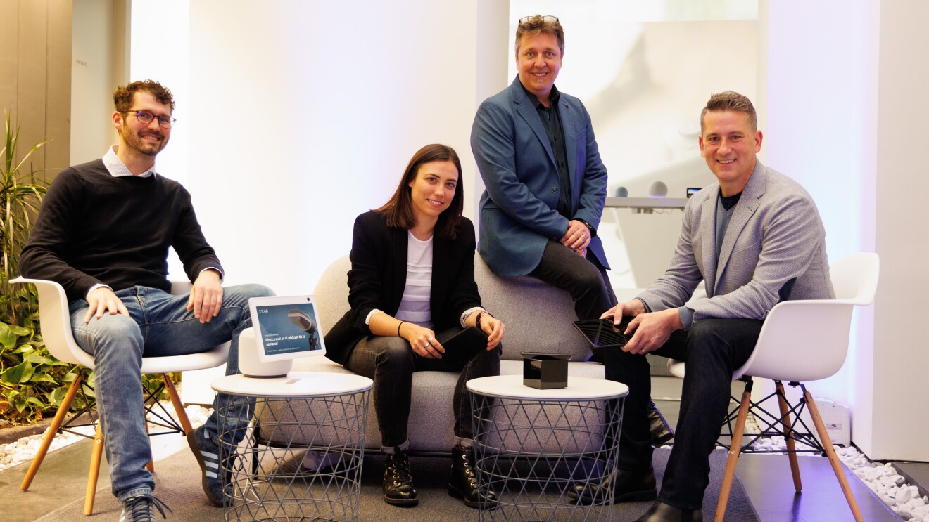 Equipo de Amazon Lab 126 en España. En la foto, el interior de un espacio de descanso de unas oficinas. Aparecen cuatro personas; tres hombres y una mujer. Están todos sentados. En prímera línea hay dos mesas donde en una hay un dispositivo de Amazon. En el centro hay un sofá de dos plazas con la mujer sentada y un hombre apoyado en el respaldo. En el límite izquierdo y derecho del sofá hay dos sillas blancas con las otras dos personas sentadas.   Descripción de todos las personas que aparecen de izquierda a derecha. El primero lleva unas deportivas, unos jeans, una camisa con un jersey, gafas y el pelo corto y barba de dos días. La mujer va con unas botas, un patalón y una americana negra. Debajo lleva una camiseta blanca. El pelo largo marrón. El segundo hombre lleva pantalones negros y americana y camisa azul oscura. Tiene perilla blanca, pelo entre rubio y blanco y las gafas en la cabeza. El tercer hombre lleva botas y pantalón negro, una americana de color gris y un jersey de punto debajo de color azul. El pelo corto y rubio. En la mano él y la mujer están sujetando un dispositivo Amazon. 