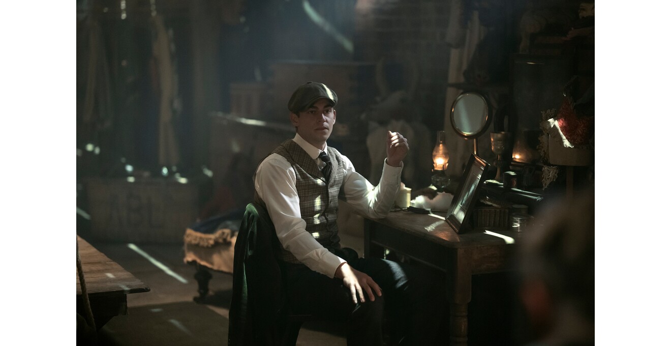 細長い隙間からの光が複数差し込む、薄暗い屋内。ハンチング帽とベストの男性が、机に向かって座っている。机の上に額縁と丸井鏡。