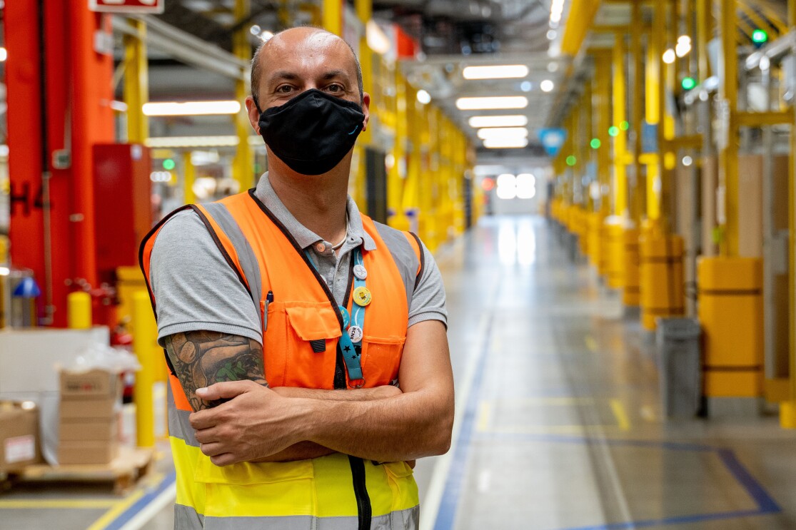 Oreste Tagliaferri, director general de los centros de distribución de España e Italia. En un pasillo del centro logístico está con los brazos cruzados Oreste. El pasillo tiene estanterías metálicas de color amarillo. Oreste tiene el brazo derecho tatuado. tiene el pelo muy corto y la parte superior no tiene pelo. Lleva una mascarilla de Amazon de color negro, un chaleco de color naranja y un polo de color gris.