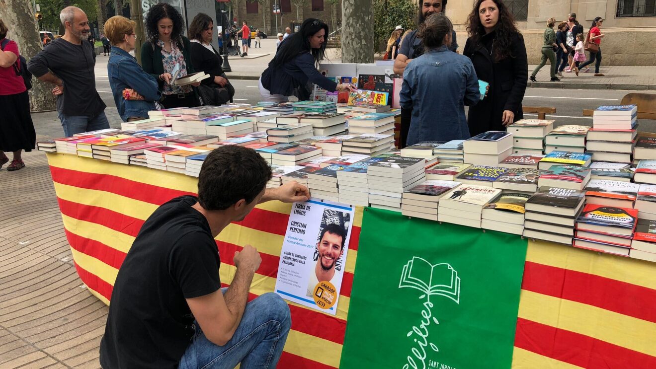 Cristian Perfumo, el escritor de la Patagonia, está de rodillas enganchando un cartel promocional de la firma de sus libros para Sant Jordi en una mesa llena de libros también suyos.
