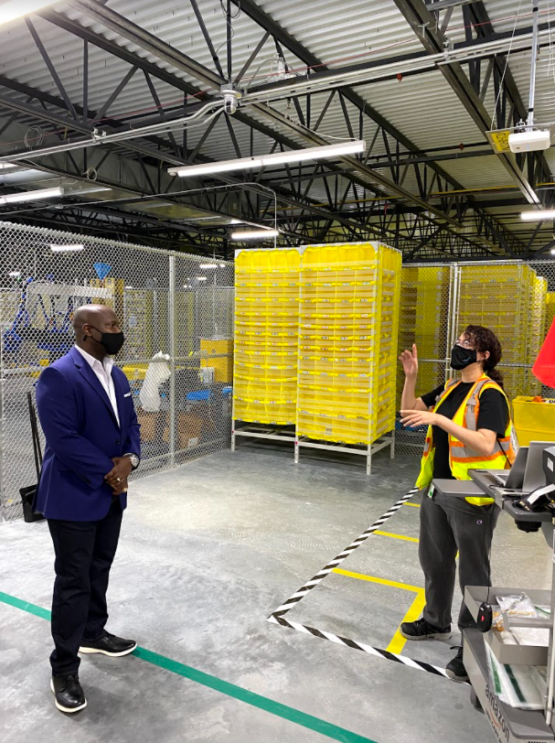 A Black man and a woman stand in an Amazon fulfillment center and engage in conversation. Both wear a face mask.