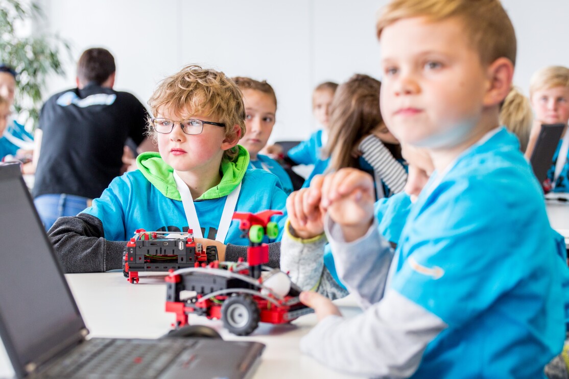 Auf einem Tisch steht ein Laptop, vor dem ein Junge im blauen T-Shirt sitzt. Im Hintergrund ist ein weiterer Junge zu sehen. Beide haben vor sich einen Fischertechnik-Transportroboter.