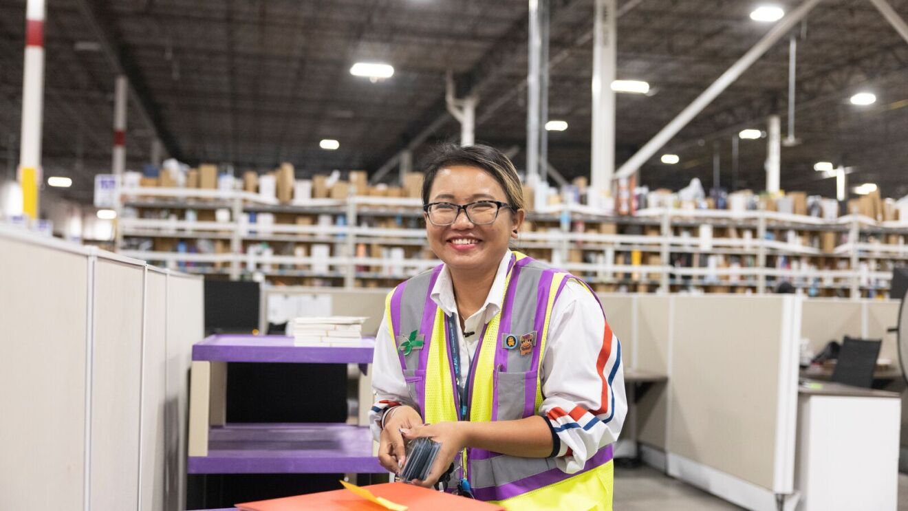 woman wearing glasses and a safety vest holding a stack of badges and smiling inside an amazon fulfillment center