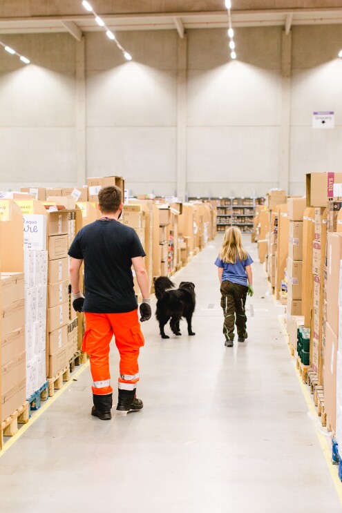 Ein Mädchen und ein Mitarbeiter der Rettungshundestaffel laufen durch den Palettengang eines Logistikzentrums, vor ihnen: ein schwarzhaariger Rettungshund.