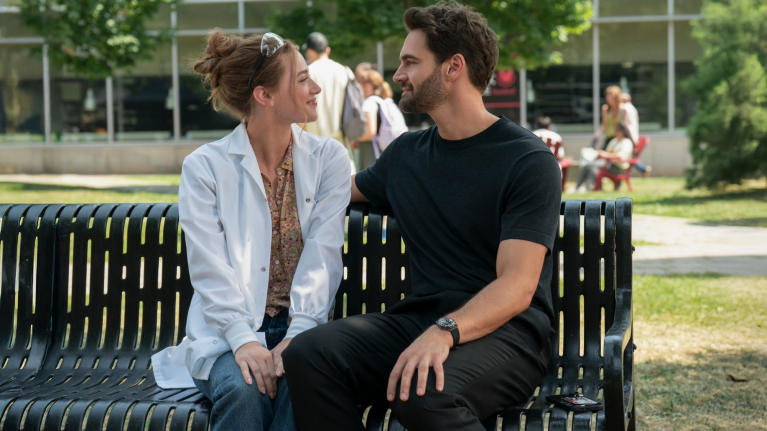 Tom Bateman and Lili Reinhart sitting on a bench in the park looking into each other's eyes
