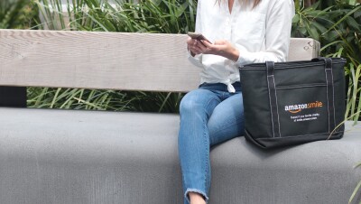 一个坐在长凳上的女人拿着智能手机。她坐在一个印有AmazonSmile标志的手提包旁边，上面写着“在smile.amazon.com上支持你最喜欢的慈善机构”。