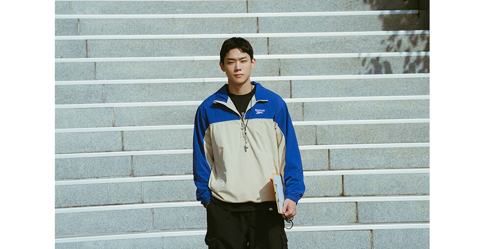 屋外で、階段を背に立つスポーツウェアの男性。左手にバインダーを持っている。