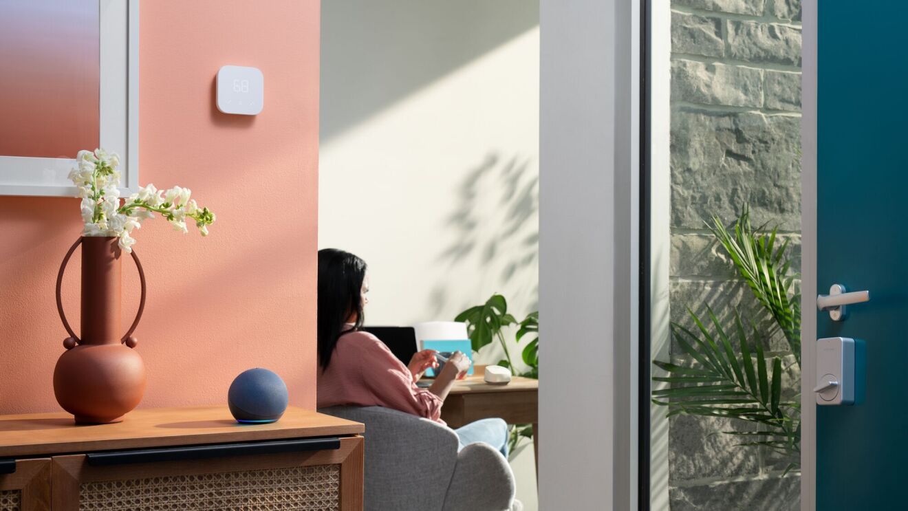 En primer plano un dispositivo Amazon y de fondo una mujer sentada delante de una mesa.