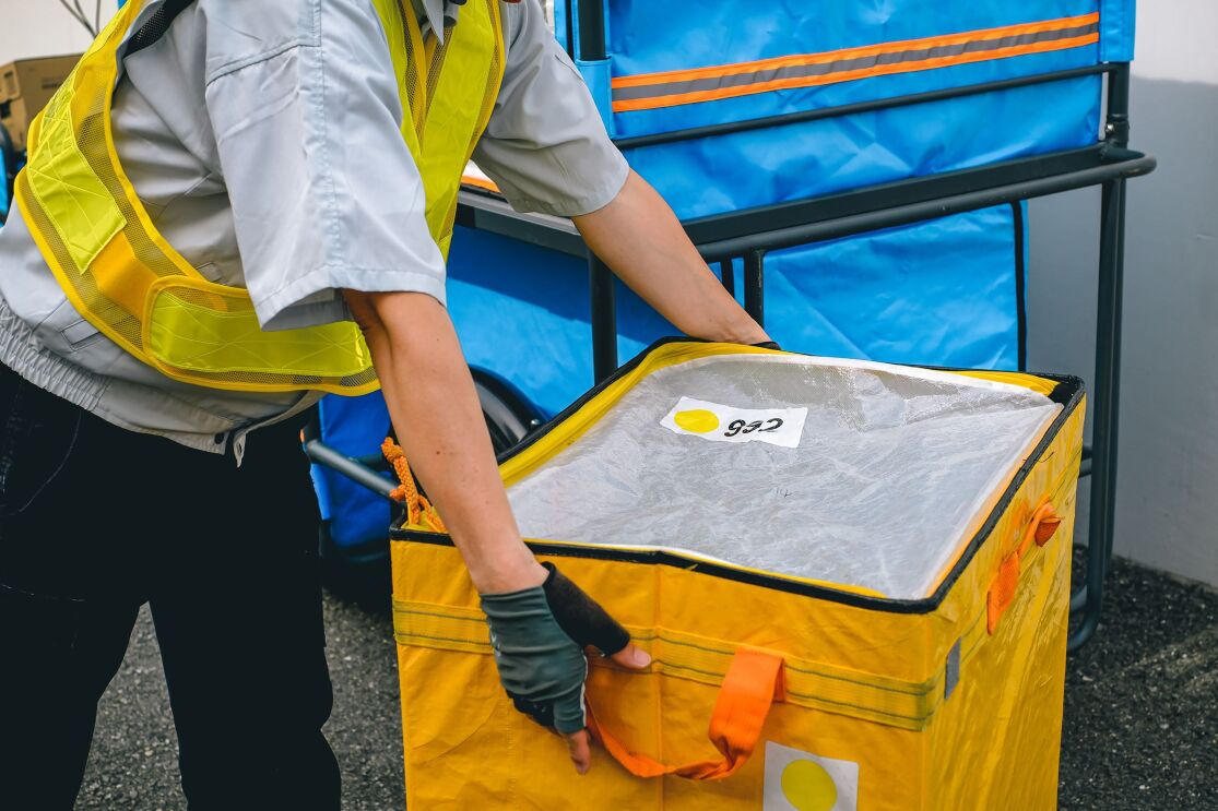 黄色い安全用ベストを着た人が緑のグローブをつけて大きな黄色い箱を運んでいる。その人の後ろには青いカバーが付いたリヤカーが置かれている。