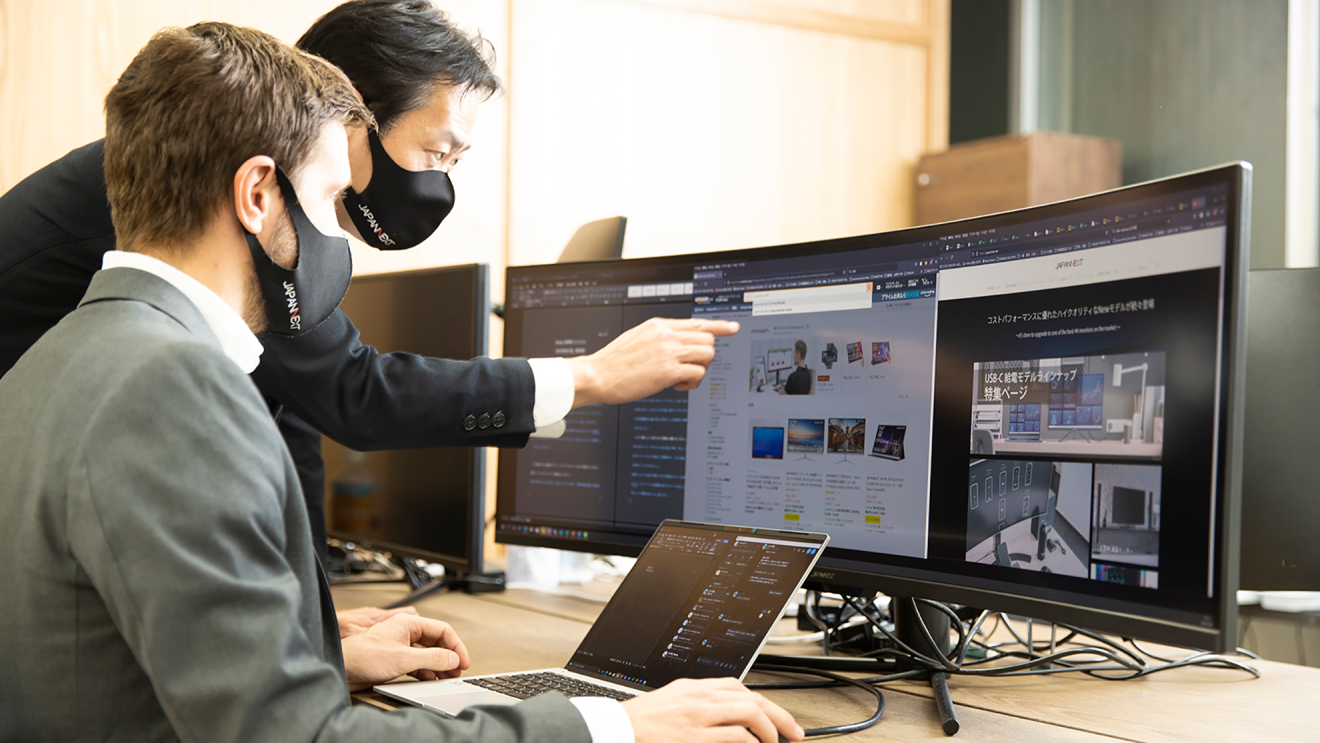 パソコンの前に座るサムエルさんと、その隣に立ちモニターを指さす剣持さんが相談している様子