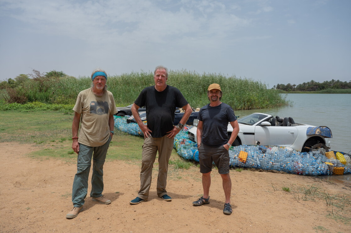 Three older men stand next to a body of water and three cars