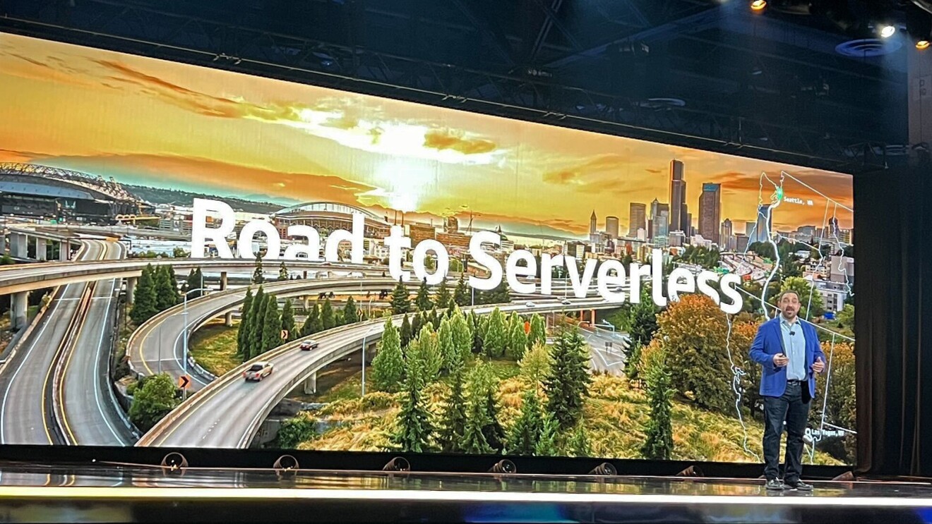 A photo of Peter DeSantis, senior vice president of AWS Utility Computing on stage at AWS re:Invent 2023. In the background is a graphic that shows the Seattle skyline at sunset. In the forefront is text that states, "Road to Serverless".