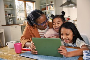 Mother and two daughters laugh at kitchen table while viewing content on an Amazon tablet. 