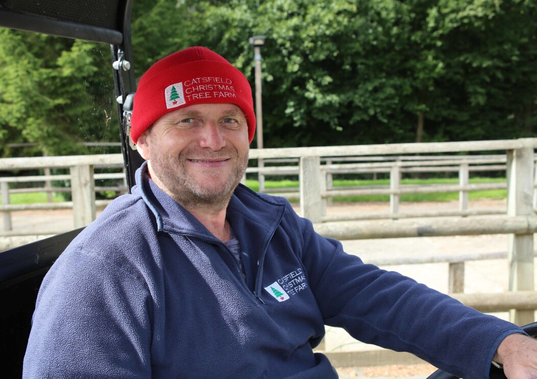 Clive Collin, owner of Catsfield Christmas Tree Farm, sitting in a buggy outside of his Christmas tree farm. He is wearing a beanie hat with his company logo on it.