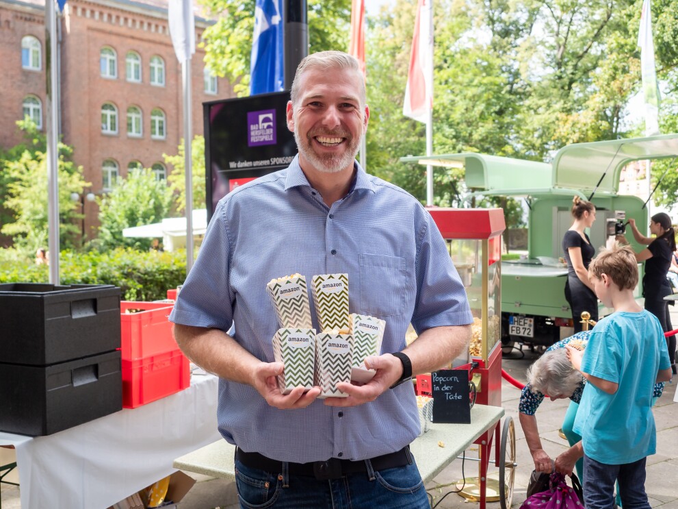 Ein Mann in Jeans und blauem Kurzarmhemd hält Popcorn-Tüten mit Amazon Aufdruck in der Hand.