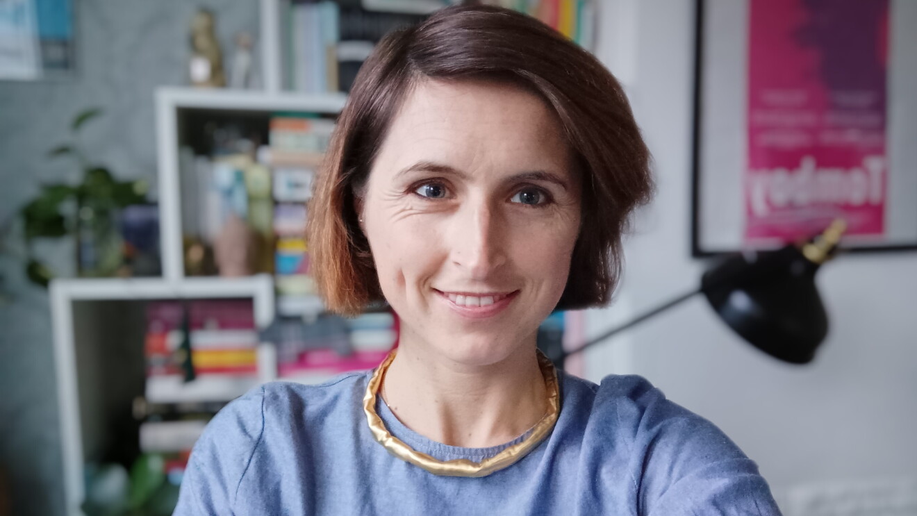Una donna con una camicia blu, i capelli tagliati sopra le spalle, una collana dorata e gli occhi chiari sorride guardando l'obiettivo. Sullo sfondo, una libreria, una lampada e alcune piante. 
