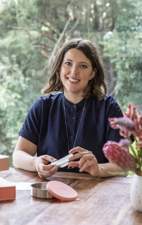 A woman sitting at a table looks to the camera and smiles.