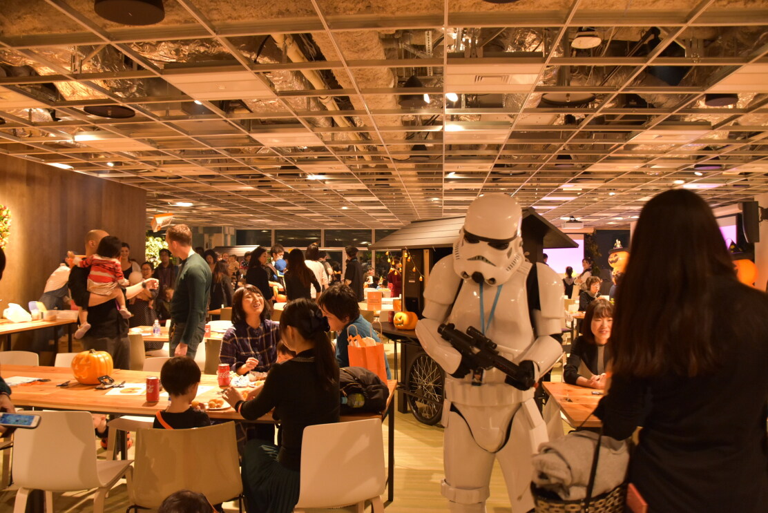 広いカフェスペースで食事や会話を楽しむ人々。中央の人物は白いアーマースーツを着用し、モデルガンを持っている。