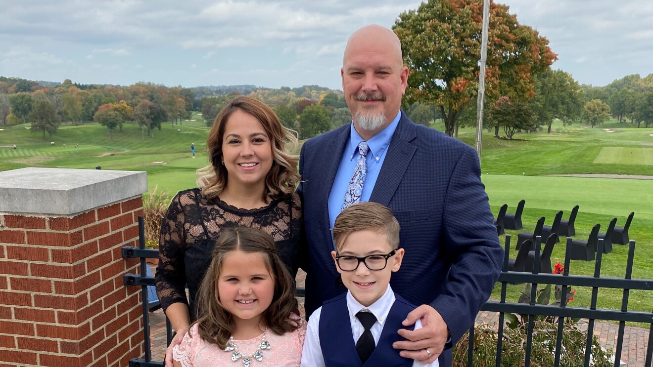A family of four stand on a pavilion, dressed up, and smile at the camera.