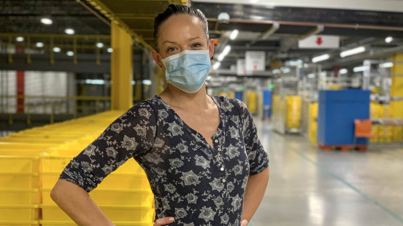 Una asociada está dentro del centro logístico. Lleva mascarila azu l, un jersey azul estampado con unoz dibujos de color blanco. Las mano las tiene en la cintura. De fondo cajas de color amarillo y azul.  