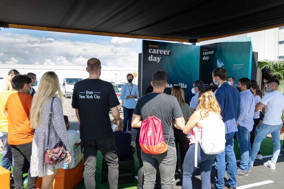 En España, el Career Day contó con una parte virtual y otra presencial, que se celebró en el nuevo centro logístico de Amazon en Illescas (Toledo). En primer plano un grupo de 20 personas de pie y se ve también que hay gente sentada en unos sillones de color naranja. Todas ellas de espaldas. Les habla una persona que está de pie y que se ve gesticular con mascarilla. De fondo el centro logístico de Amazon en Illescas. La gente está escuchando y en la parte derecha hay un cartel que llega al suelo y pone Amazon Career Day y Úlitma milla. Se ve el cielo azul con nubes blancas. 