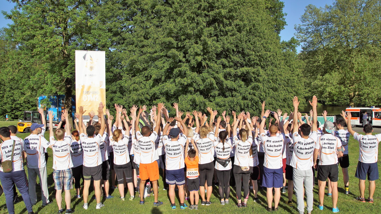Eine Gruppe von Läufern in kurzen Hosen und in Amazon Shirts hat sich auf einer Wiese versammelt. Sie sind von hinten zu sehen, alle strecken die Hände in die Höhe. Auf den Shirt-Rücken steht: "Mit einem Lächeln ins Ziel".