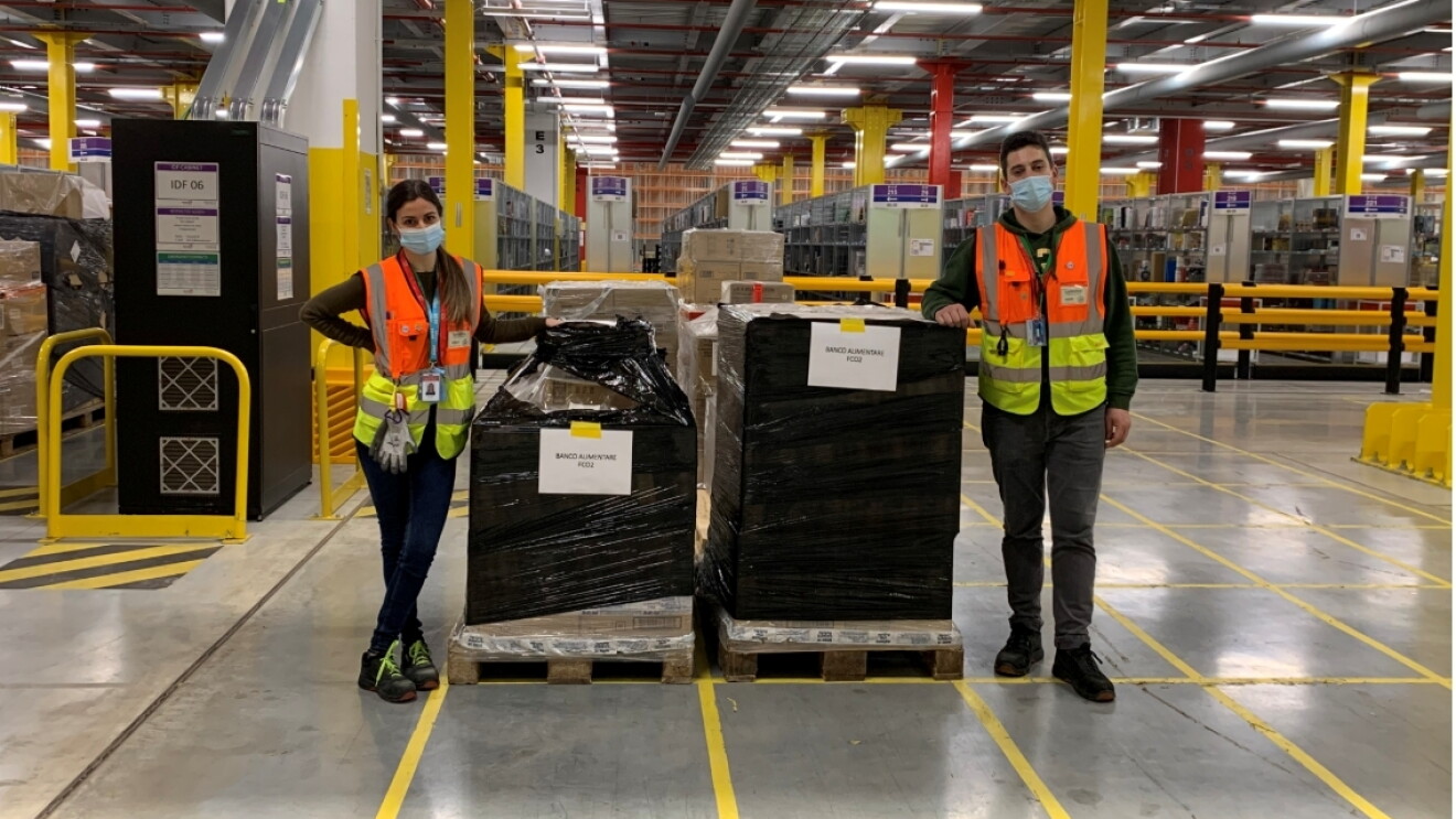 Due dipendenti Amazon, un uomo e una donna, posano accanto a due pallet di materiale destinato a Banco Alimentare. La donna posa sulla sinistra, mentre l'uomo sulla destra. Il materiale sui pallet è ricoperto da una pellicola nera. Entrambi indossano giubbini catarinfrangenti arancioni e gialli e una mascherina chirurgica sul volto. Si trovano all'interno di un magazzino Amazon e sono ripresi a figura intera. 