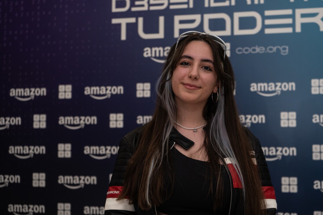 Con un fondo de photocall con el logo de Amazon y de Code.org, en blanco sobre un fondo azul aparece unos de los finalistas, Irene Verdesoto Sánchez. Lleva el pelo muy largo, por debajo del pecho, es liso y tiene una mecha a cada lado de color blanco. Va con una cazadora negra, amarilla y blanca y un jersey debajo de color negro. Lleva un collar pendientes de aros y las gafas de sol apoyadas en el pelo.