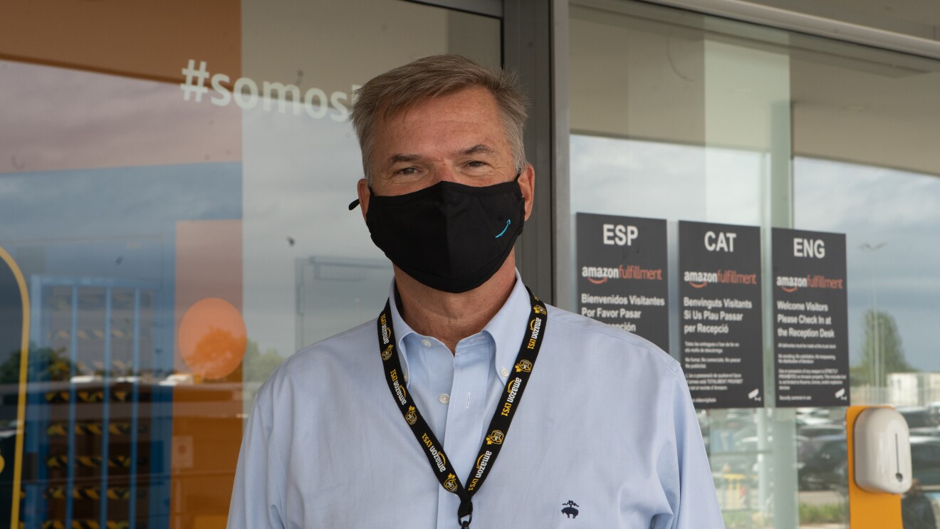 Fred Pattje, Director de Amazon Customer Fulfillment en Francia, Italia y España. De fondo el exterior de un centro logístico con unos carteles negros y letra blanca en español, catalán e inglés. También un dispensador de gel y una puerta de color azul. El protagonista va con una mascarilla corporativa de Amazon. Una camisa de color azul. Está moreno y el pelo es castaño y blanco con los ojos marrones.