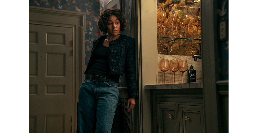 ジーンズ姿の巻き毛の女性が花柄の壁紙の壁にもたれている。女性がもたれた壁の横にはワイングラスが多数並んだ棚が見える。