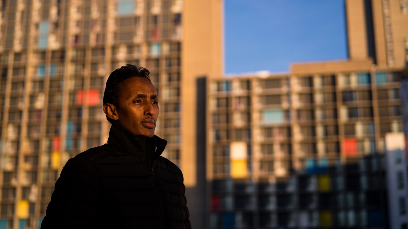 A man is photographed from the torso up standing in front of large buildings. The buildings are interspersed with bright rectangles of color in place of some of the windows.