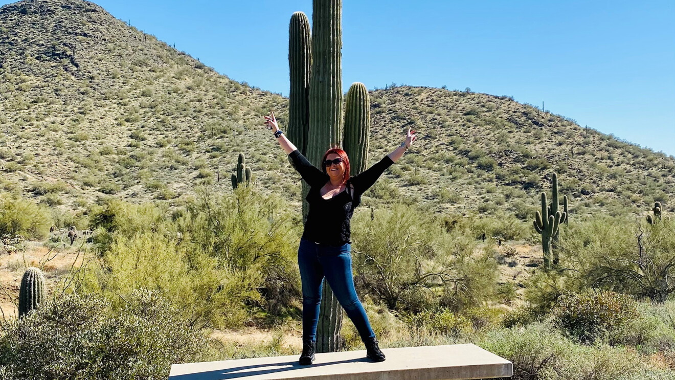 An image of a woman standing in front of a cactus with her arms in the air posing for a photo. 