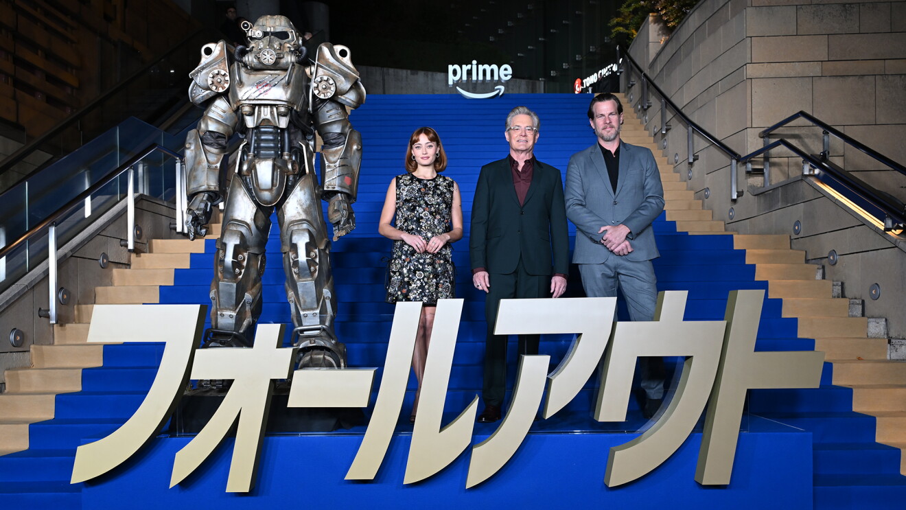 青いカーペットが敷かれた大階段に立つ3人の人物。脚の前に「フォールアウト」の文字のオブジェが置かれている。3人の左側にパワーアーマースーツ。
