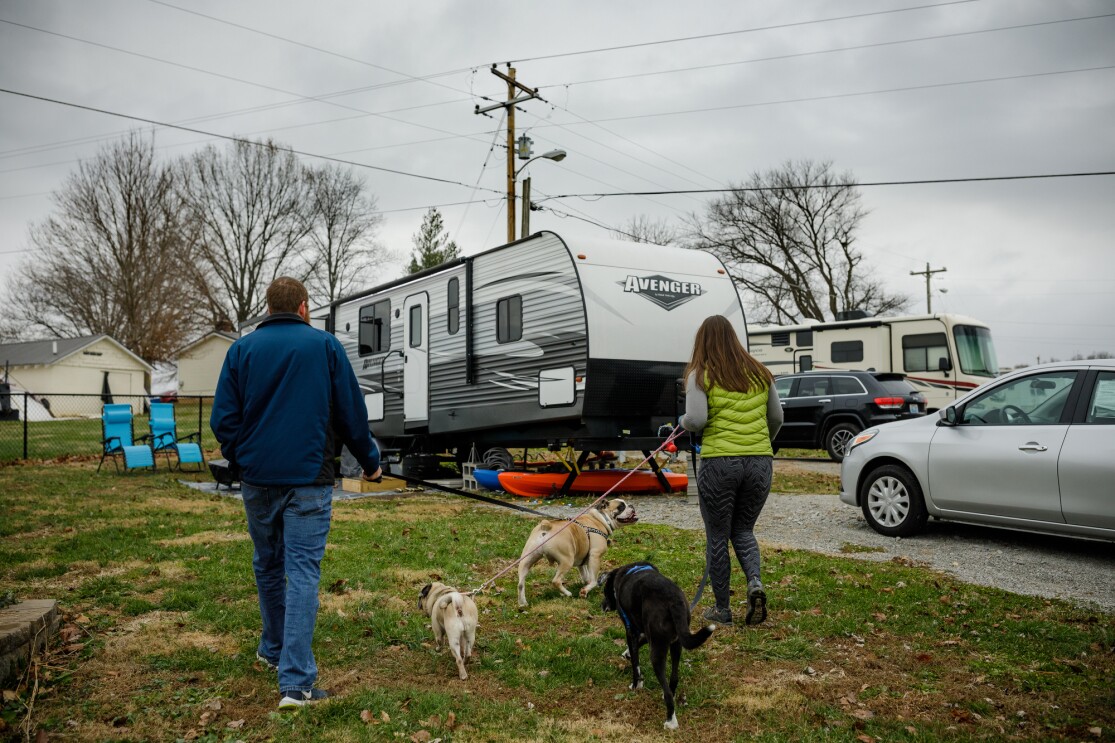 A man, a woman, and three leashed dogs walk toward a parked RV. 