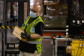A man wearing gloves, a mask, and a safety vest carries a small box.
