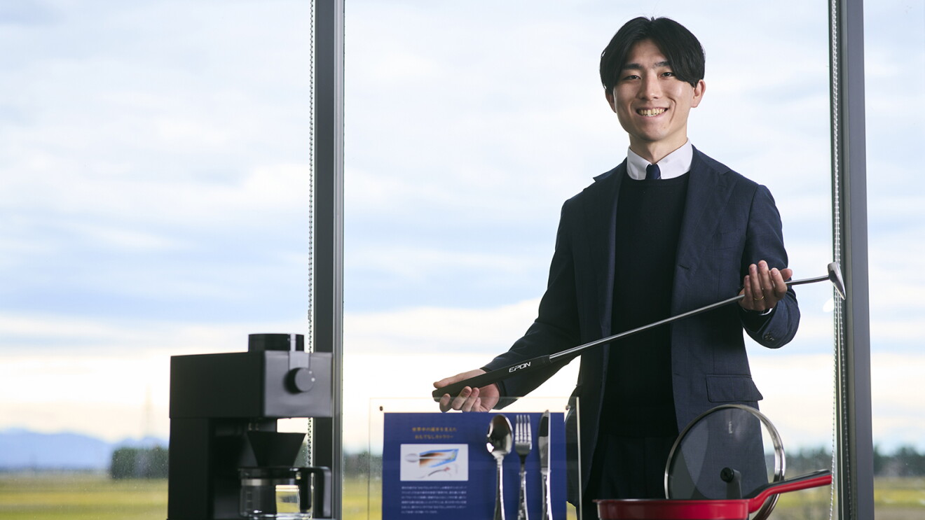 大きな窓を背に立つスーツ姿の男性。両手でゴルフのパターを持っている。男性の前側にコーヒーマシンやカトラリーサンプル、フライパンと蓋。