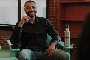 Smiling man in black shirt seated in teal chair with Amazon water bottle nearby